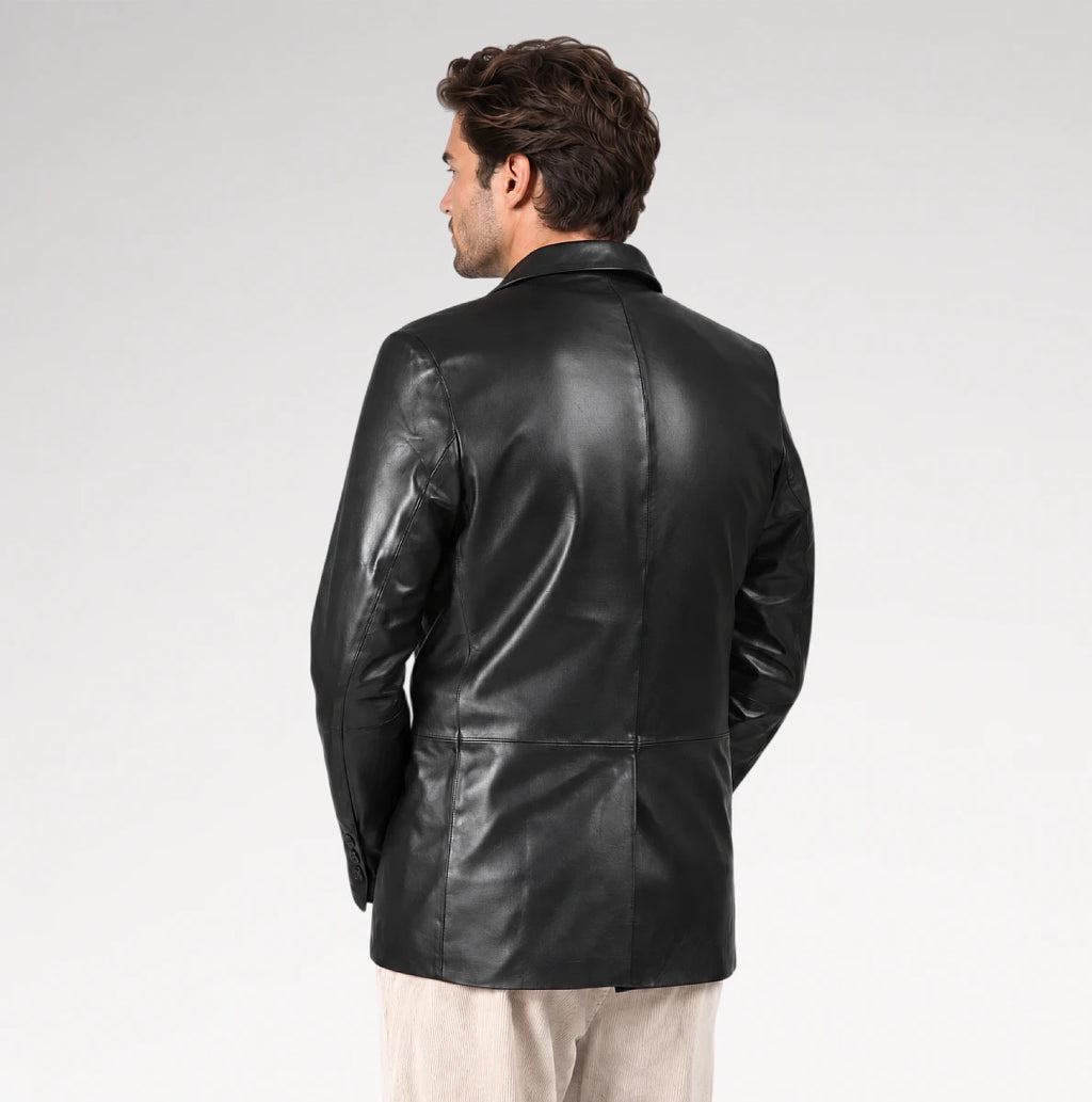 A man viewed from the back wearing a black leather blazer over a light beige shirt and trousers, set against a plain light gray studio background.