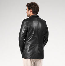 A man viewed from the back wearing a black leather blazer over a light beige shirt and trousers, set against a plain light gray studio background.