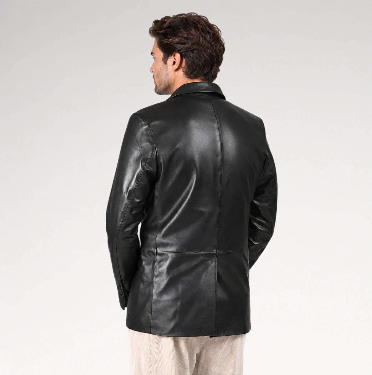 A man viewed from the back wearing a black leather blazer over a light beige shirt and trousers, set against a plain light gray studio background.