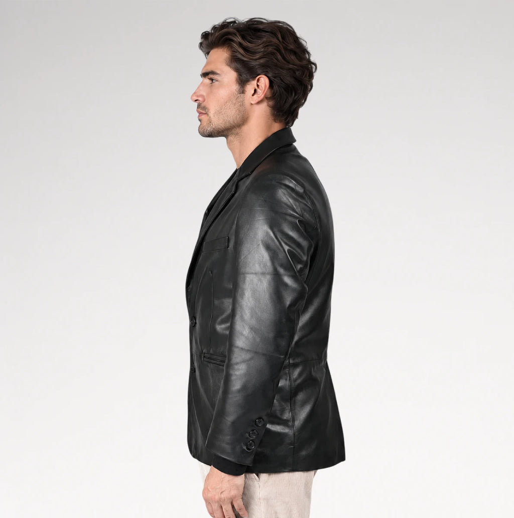 A man in profile wearing a black leather blazer over a dark shirt and light beige trousers, photographed against a clean light gray studio background.