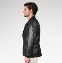 A man in profile wearing a black leather blazer over a dark shirt and light beige trousers, photographed against a clean light gray studio background.