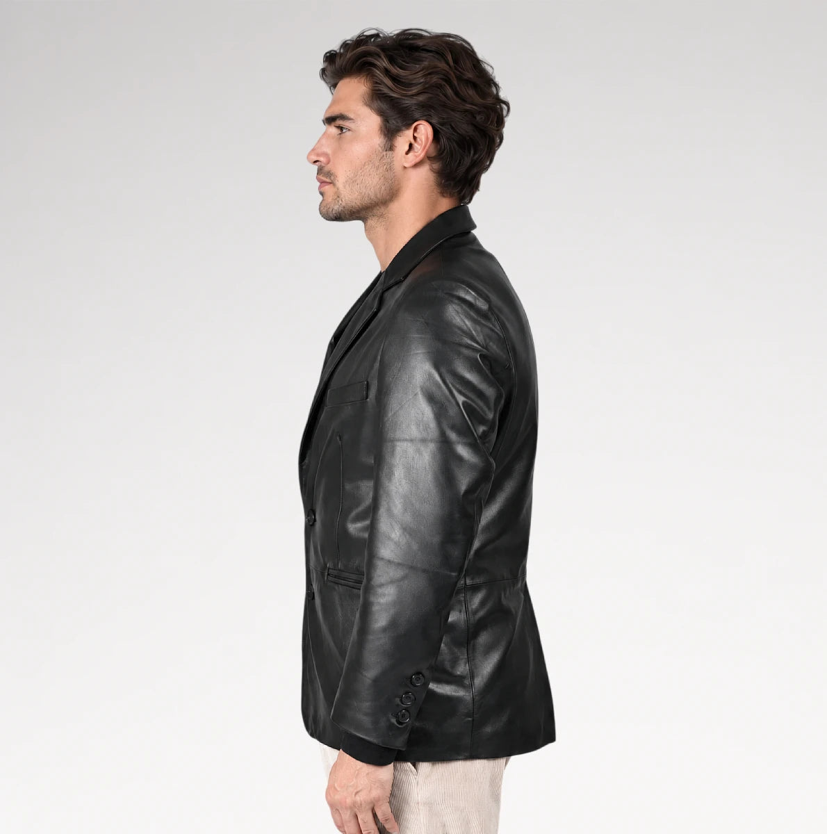 A man in profile wearing a black leather blazer over a dark shirt and light beige trousers, photographed against a clean light gray studio background.