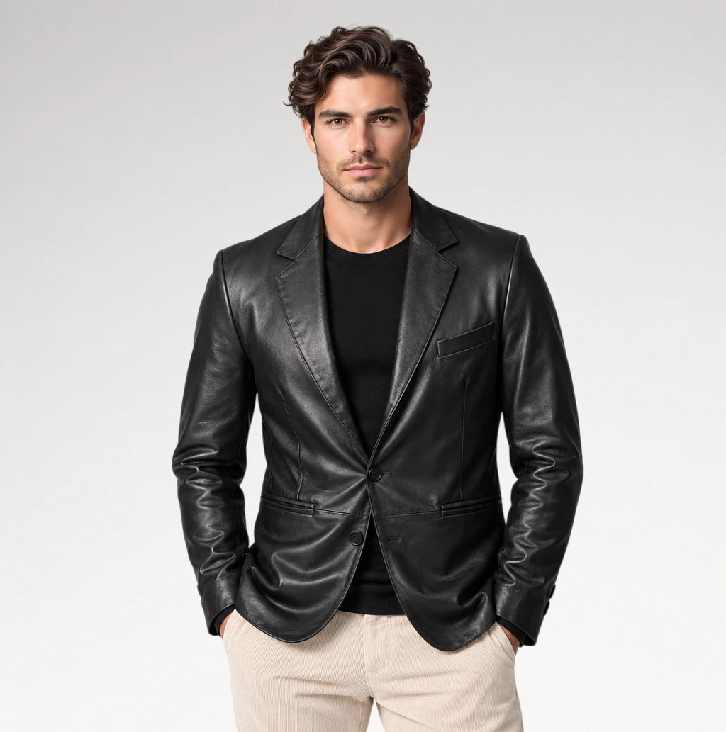 A man wearing a black leather blazer over a black t-shirt and beige trousers, standing against a light gray studio background.