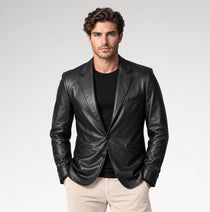 A man wearing a black leather blazer over a black t-shirt and beige trousers, standing against a light gray studio background.