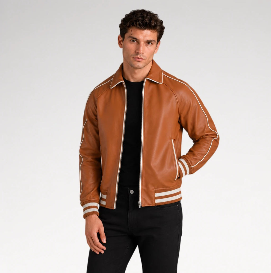 A man wearing a brown leather varsity-style jacket with white contrast piping and striped ribbed cuffs and hem, paired with a black shirt and black pants, against a light gray studio background.