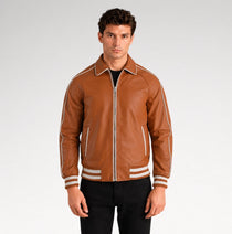 A man wearing a brown leather varsity-style jacket with white contrast piping and striped ribbed cuffs, paired with black pants, against a light gray studio background.