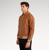 A man wearing a brown leather varsity-style jacket with white stripe details on the shoulders, sleeves, and cuffs, paired with black pants, photographed against a light gray studio background.