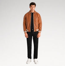 A male model wearing a brown leather varsity-style jacket with white stripes on the collar, cuffs, and hem, styled with a black t-shirt, black jeans, and white sneakers against a light gray studio background.