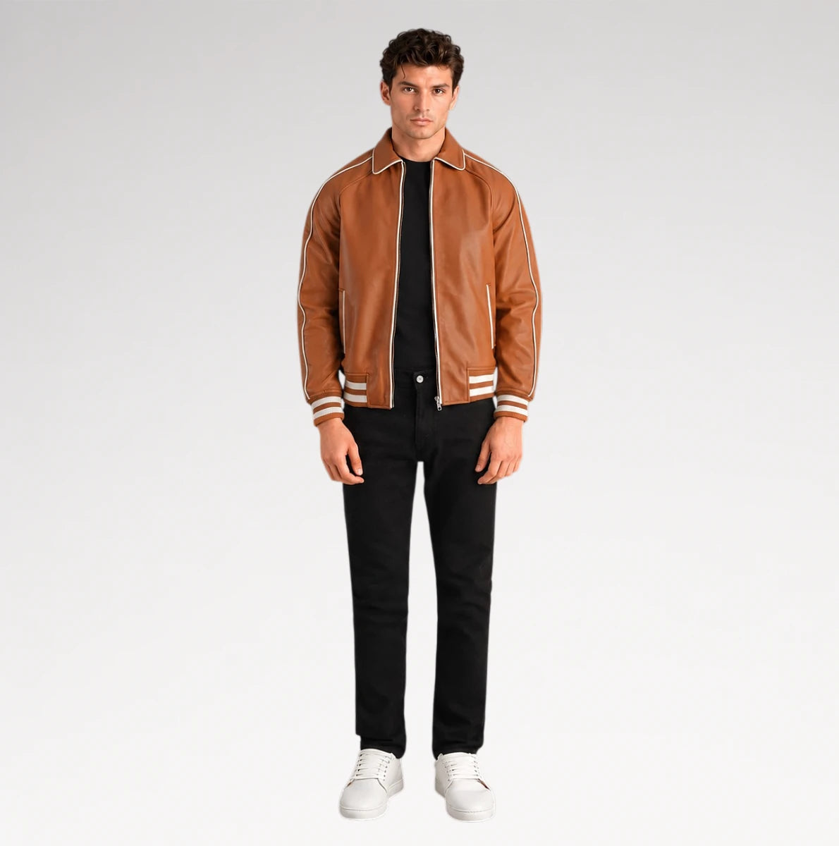 A male model wearing a brown leather varsity-style jacket with white stripes on the collar, cuffs, and hem, styled with a black t-shirt, black jeans, and white sneakers against a light gray studio background.