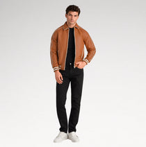 A man modeling a brown leather bomber jacket with cream stripes on the shoulders and cuffs, paired with a black t-shirt, black pants, and white sneakers, against a light gray studio background.