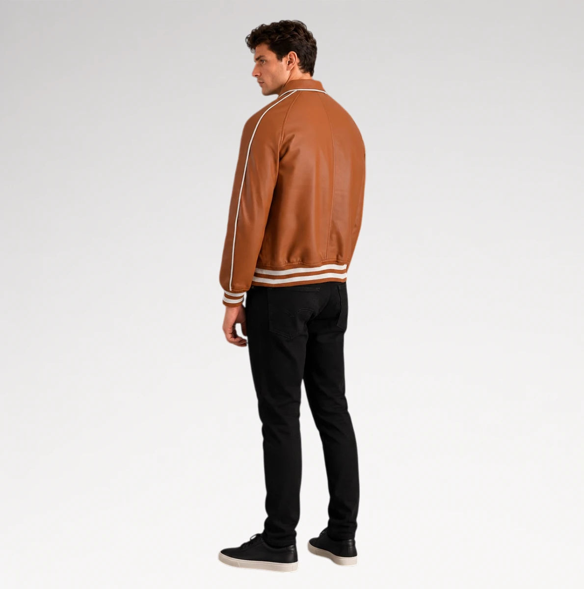 A man wearing a brown leather varsity-style jacket with white stripes on the shoulders and hem, paired with black pants and black sneakers, shown from a rear three-quarter view against a light gray studio background.