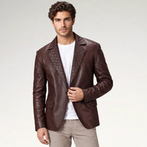 A man wearing a dark brown leather blazer with quilted lapels and pocket details, layered over a white t-shirt and beige trousers, photographed against a light gray studio background.
