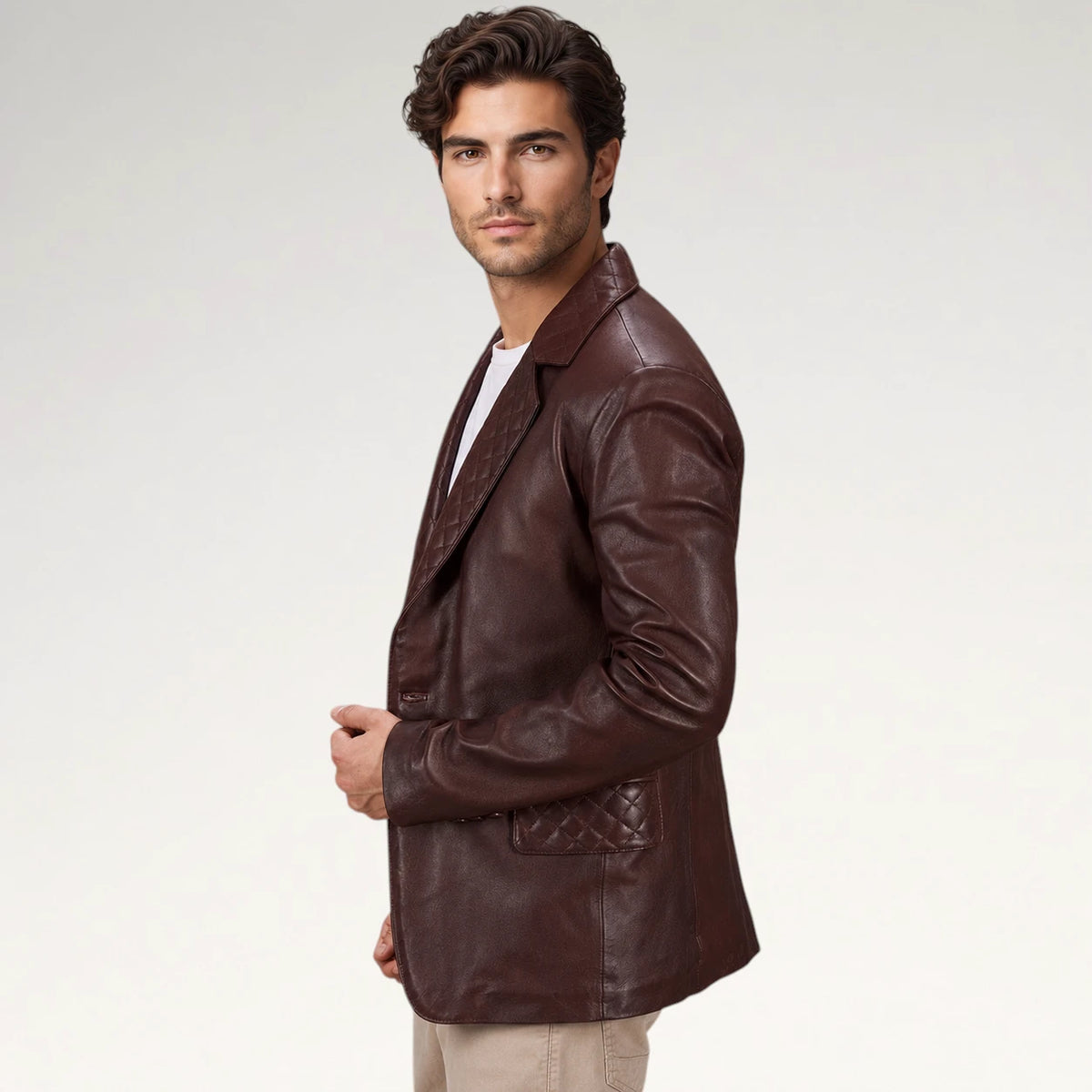 A man wearing a dark brown leather blazer with a quilted pocket detail, styled over a white t-shirt and beige pants, photographed against a light gray studio background.