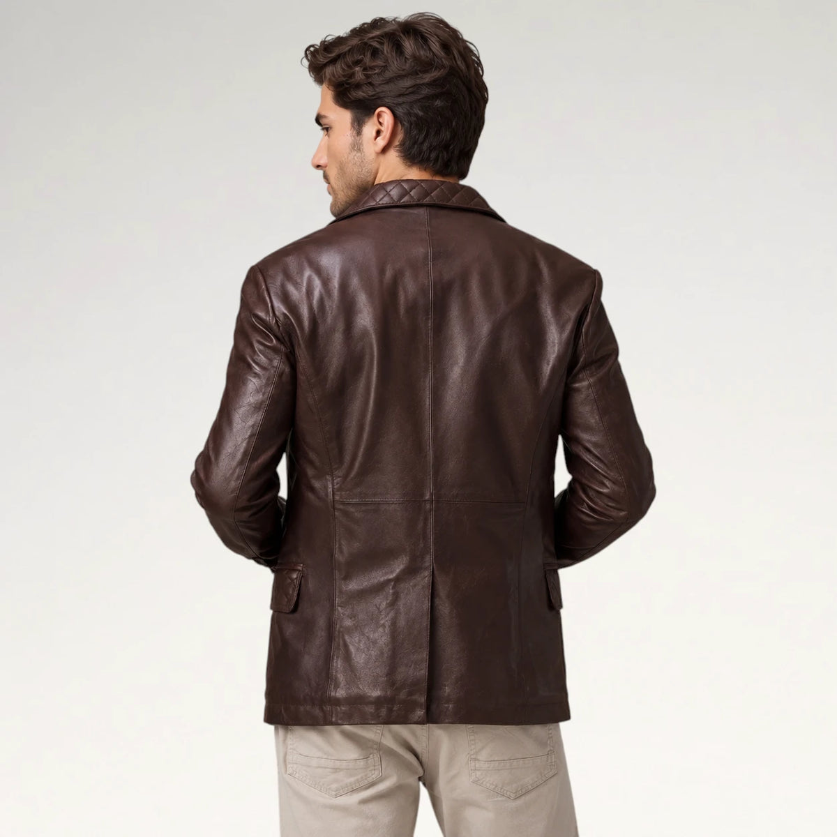 A man viewed from the back wearing a dark brown leather blazer with a quilted collar and beige trousers, set against a plain light gray studio background.