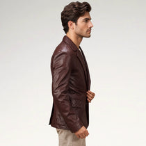 A man in profile wearing a dark brown leather blazer with quilted details on the chest and waist, paired with beige trousers, against a plain light gray studio background.