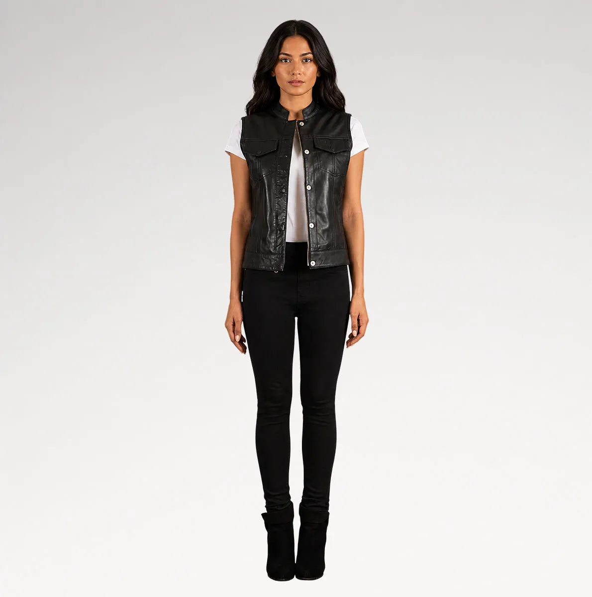 A woman modeling a black leather sleeveless vest over a white t-shirt and black skinny jeans, standing against a light gray studio background.