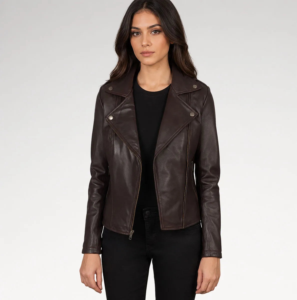 A woman modeling a dark brown faux-leather moto jacket with silver snap and zip details, worn over a black top and black pants against a light gray studio background.