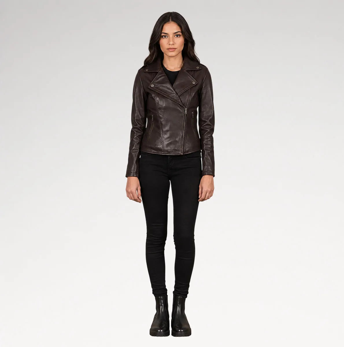 A woman modeling a dark brown leather moto jacket, paired with black skinny jeans and black ankle boots, against a light gray studio background.