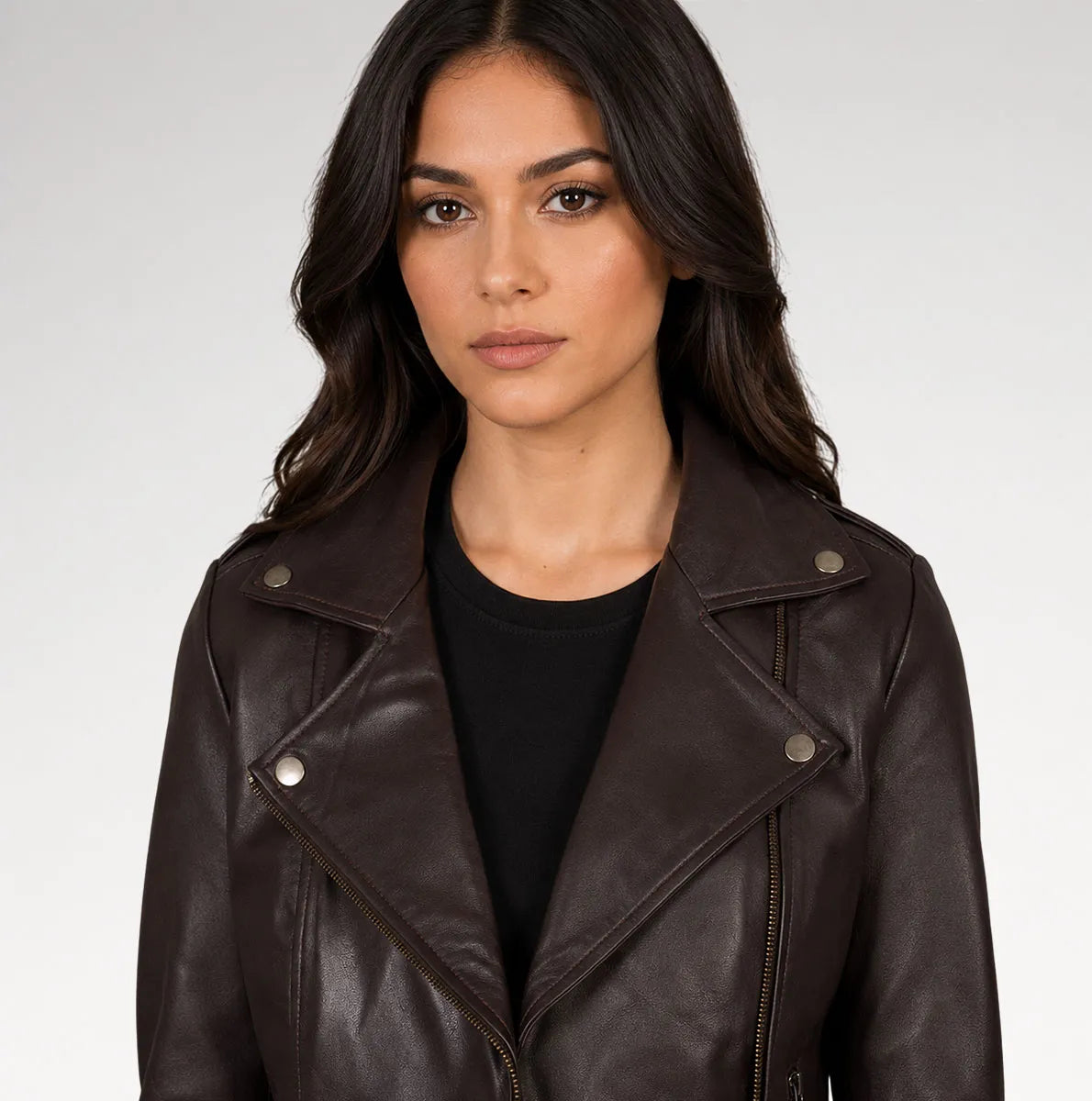 A woman wearing a dark brown faux-leather moto jacket over a black top, photographed against a light gray studio background.