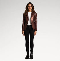 A woman stands against a light gray studio background wearing a metallic dark brown leather jacket over a white top, black skinny pants, and black ankle boots.