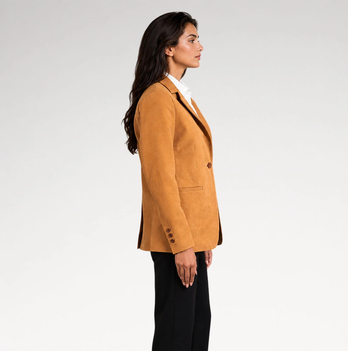 A woman shown in side profile wearing a camel-colored suede blazer over a white collared shirt and black trousers, photographed against a clean light gray studio background.