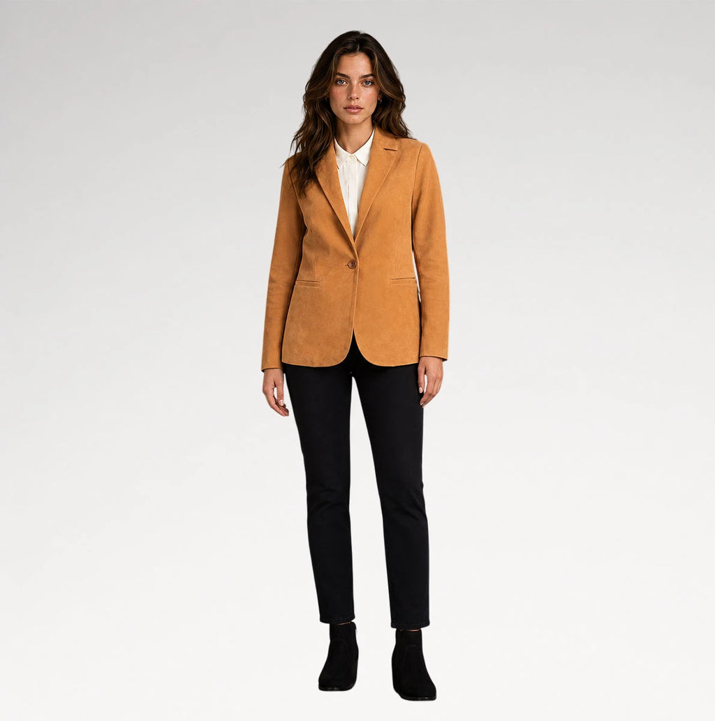 A woman modeling a camel-colored suede blazer over a white collared shirt, paired with black trousers and black ankle boots, against a light gray studio background.