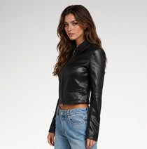 A woman modeling a black faux-leather cropped jacket with a collar and front zipper, paired with light-wash blue jeans, against a plain light gray studio background.