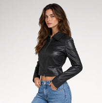 A woman modeling a black cropped faux-leather jacket with a collar and front zipper, paired with blue jeans, against a light gray studio background.