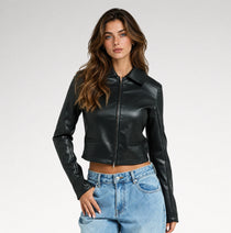 A woman modeling a black faux-leather cropped jacket with a front zipper and collar, paired with light blue jeans, against a plain light gray studio background.