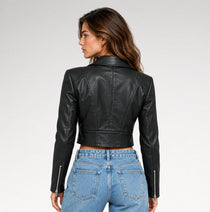A woman shown from the back wearing a black cropped faux-leather moto jacket with silver zipper details on the sleeves, paired with light blue jeans, against a light gray studio background.
