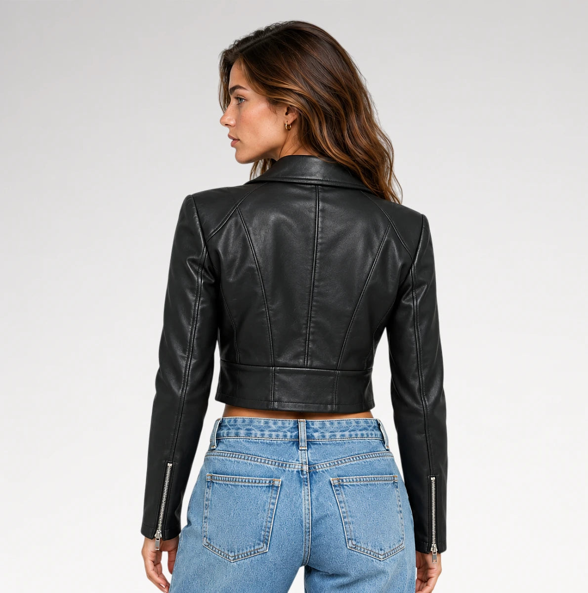 A woman shown from the back wearing a black cropped faux-leather moto jacket with silver zipper details on the sleeves, paired with light blue jeans, against a light gray studio background.