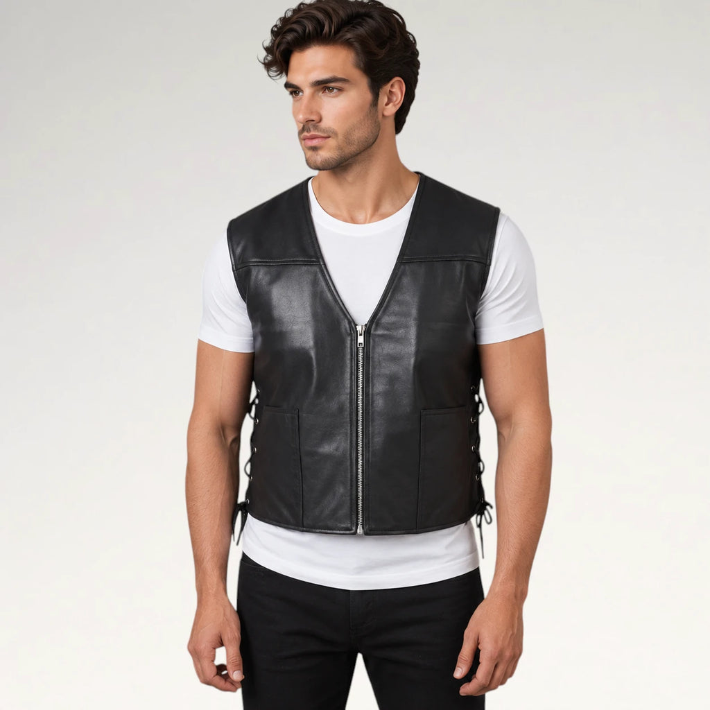 A man wearing a black leather motorcycle vest over a white t-shirt and black pants, standing against a light gray studio background.