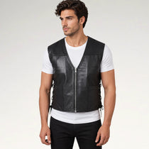 A man wearing a black leather motorcycle vest over a white t-shirt and black pants, standing against a light gray studio background.