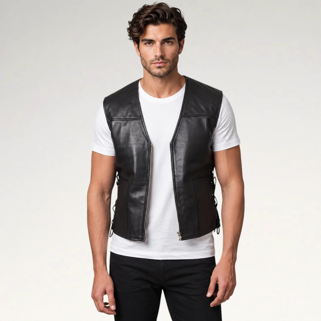 A man wearing a black leather motorcycle vest over a white t-shirt and black pants, standing against a light gray studio background.