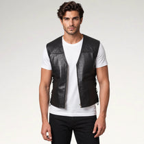 A man wearing a black leather motorcycle vest over a white t-shirt and black pants, standing against a light gray studio background.