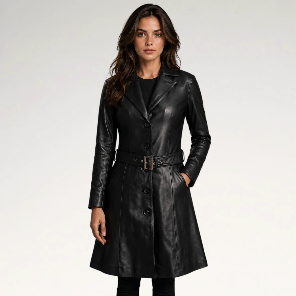 A woman modeling a black leather trench coat with a matching belt and gold-tone buckle, photographed against a clean light gray studio background.