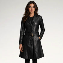 A woman modeling a black leather trench coat with a matching belt and gold-tone buckle, photographed against a clean light gray studio background.