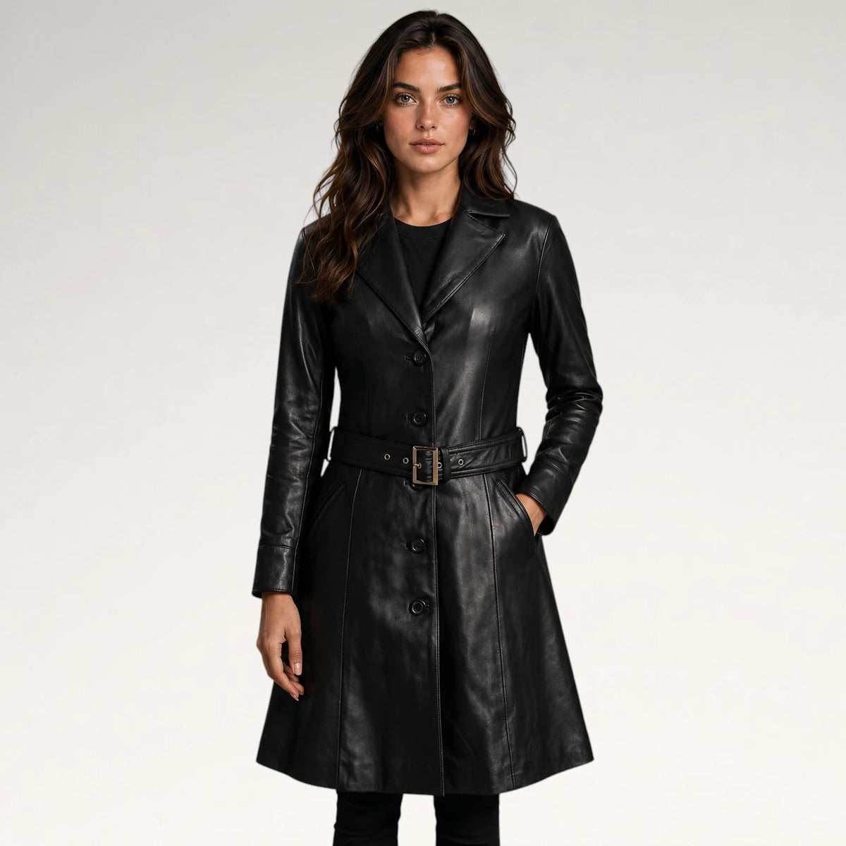A woman modeling a black leather trench coat with a matching belt and gold-tone buckle, photographed against a clean light gray studio background.