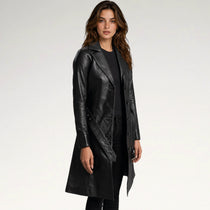 A woman modeling a black faux-leather trench coat over a black outfit, photographed against a clean white studio background.