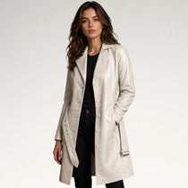 A woman modeling a light beige faux-leather double-breasted coat over a black top and black pants, photographed against a clean white studio background.