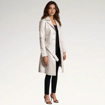 A woman modeling a long, light-colored leather coat over a black outfit, standing against a plain light gray studio background.