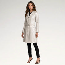 A woman modeling a light beige leather trench coat with a matching belt, paired with black pants and black high-heeled sandals, against a plain white studio background.