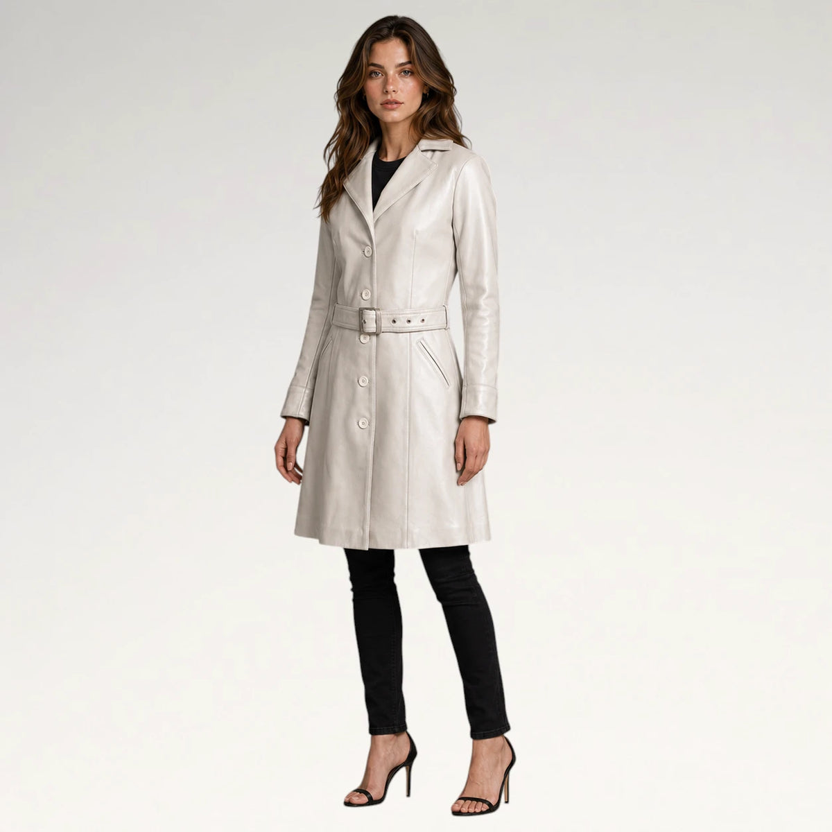 A woman modeling a light beige leather trench coat with a matching belt, paired with black pants and black high-heeled sandals, against a plain white studio background.