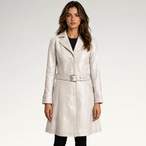 A woman modeling a light beige leather trench coat with a matching belt, worn over a black top and black pants, against a plain light gray studio background.