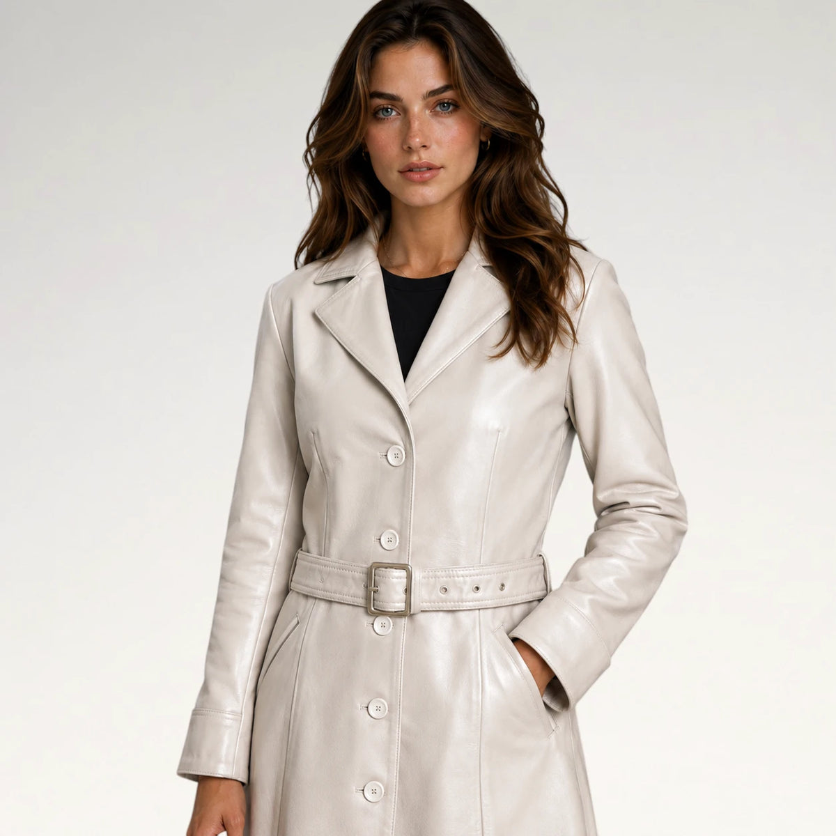 A woman modeling a light beige faux-leather trench coat with a matching belt and silver buckle, worn over a black top, against a plain light gray background.
