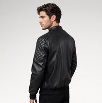 A man wearing a black leather bomber jacket with quilted shoulder panels, shown from a side-back angle against a light gray studio background.