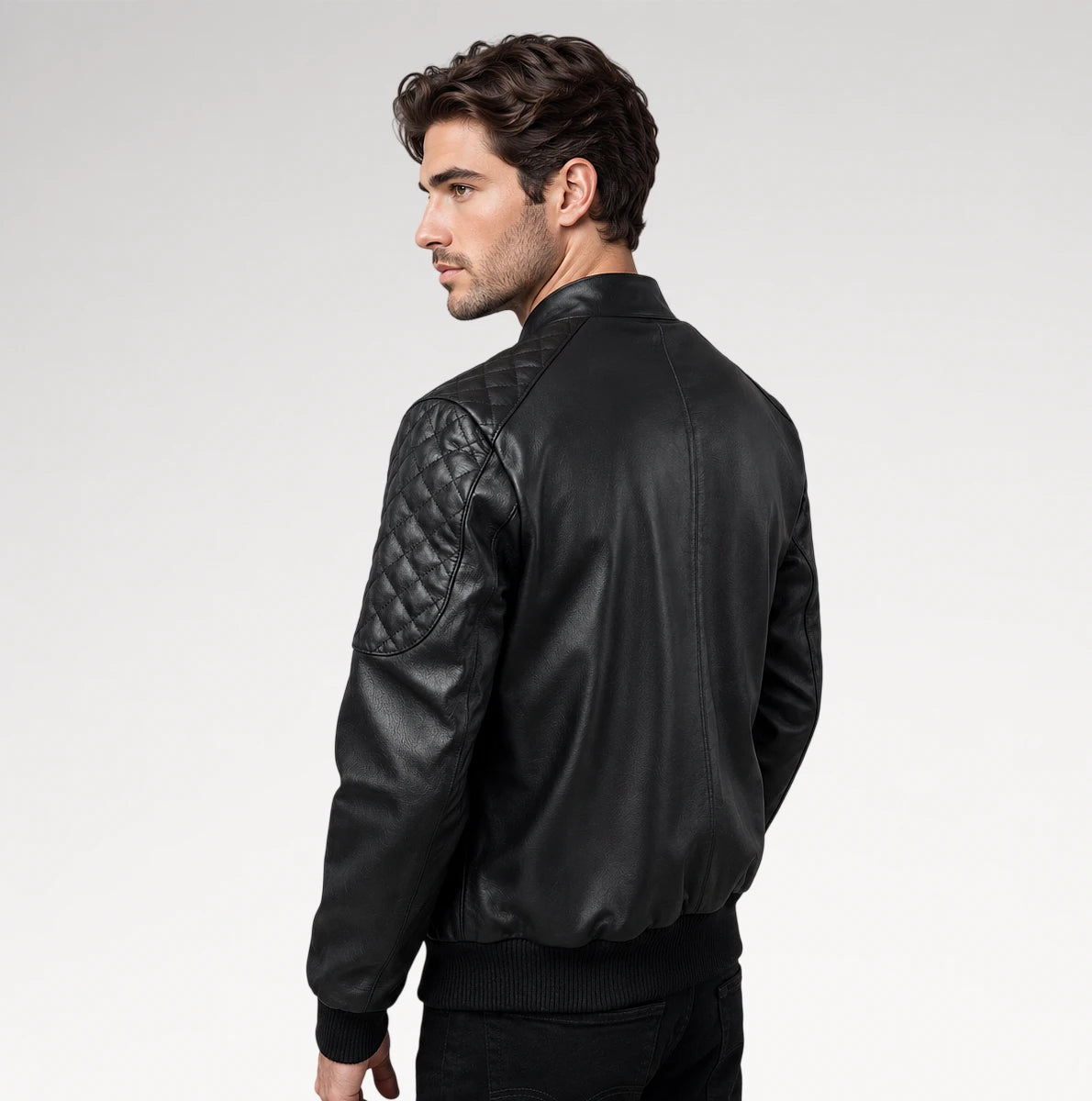 A man wearing a black leather bomber jacket with quilted shoulder panels, shown from a side-back angle against a light gray studio background.