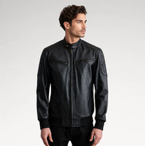 A man wearing a black leather jacket with quilted shoulder details, zippered chest pockets, and a stand-up collar, photographed against a light gray studio background.