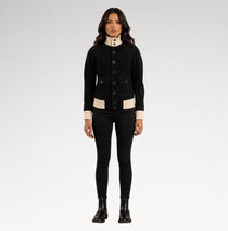 A model stands against a light gray studio background wearing a black cropped button-front jacket with beige contrast panels at the collar, cuffs, and hem, paired with black skinny pants and black ankle boots.