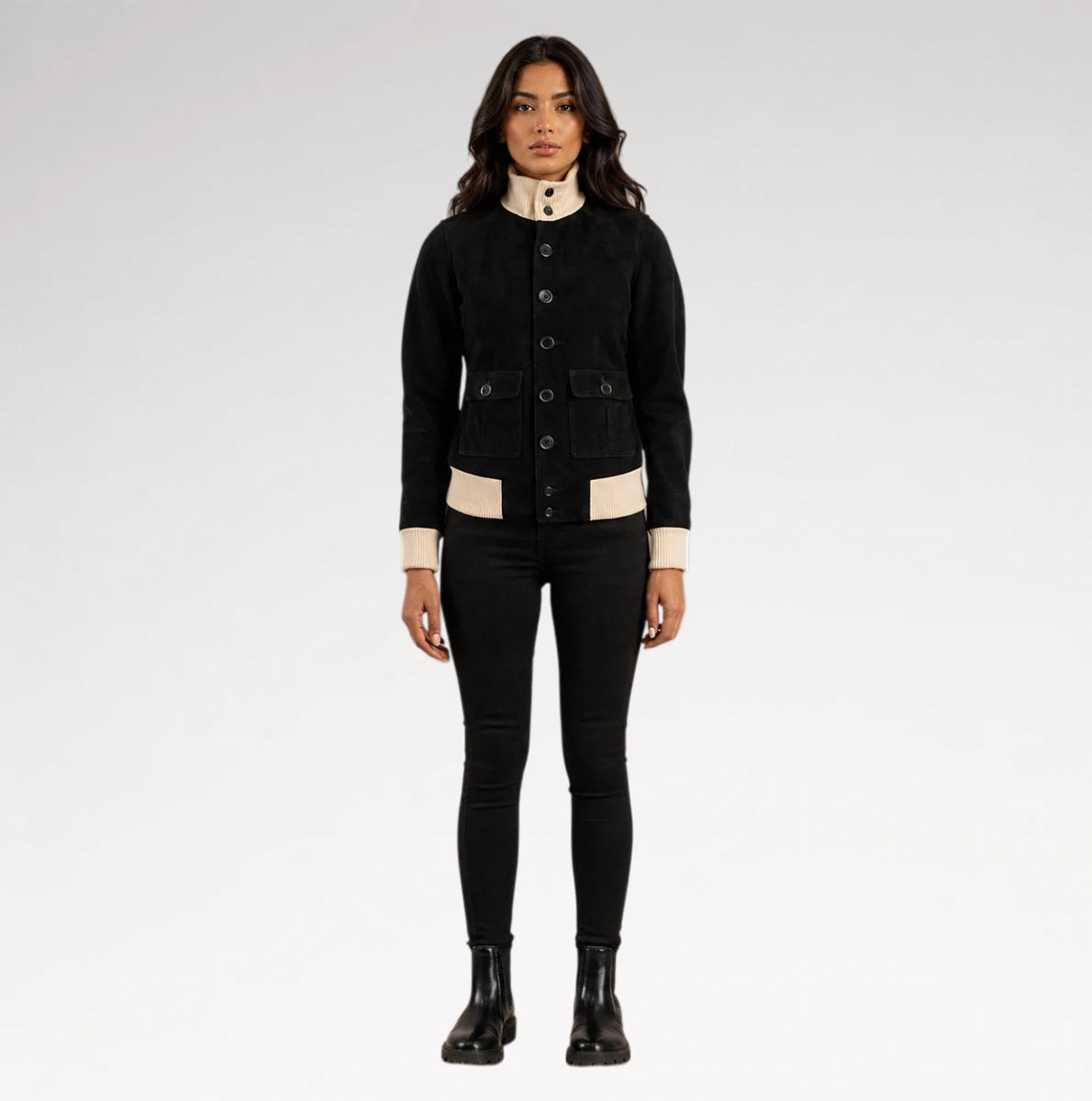 A model stands against a light gray studio background wearing a black cropped button-front jacket with beige contrast panels at the collar, cuffs, and hem, paired with black skinny pants and black ankle boots.